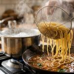 **Alt text:** Close-up of steaming spaghetti being lifted with a wire spider skimmer from boiling water into a skillet of glossy tomato sauce. Water droplets drip from the noodles, with soft steam rising in a sunlit kitchen with a shallow depth of field.