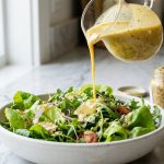 A creamy golden vinaigrette is drizzled from a glass pitcher onto a fresh green salad in a white ceramic bowl, bathed in bright, natural light.