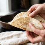 **Alt Text:** A close-up of a hand gently folding a warm, flexible flour tortilla, showcasing its soft texture and golden-brown spots as a faint wisp of steam rises.