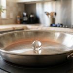 **Alt text:** A macro close-up of a single, spherical water droplet hovering and gliding on a hot, brushed stainless steel skillet, illustrating the Leidenfrost effect in a bright, modern kitchen.