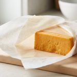 **Alt text:** A close-up, editorial photograph of a block of sharp orange cheddar cheese on a light wooden cutting board, partially unwrapped from a specialized, dual-layer white cheese paper.