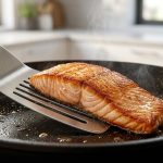 An extreme close-up of a flexible stainless steel fish spatula sliding under a perfectly seared salmon fillet in a seasoned cast-iron skillet.