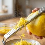 **Alt text:** An extreme macro photograph of a fresh yellow lemon being zested with a metal grater. Tiny curls of yellow zest and a fine mist of citrus oil float in the air as the grater cleanly peels the outer skin, leaving the white pith intact against a softly blurred kitchen background.