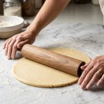 **Alt text:** Baker's hands using a wooden French tapered rolling pin to roll out smooth pastry dough on a flour-dusted marble countertop in soft light.