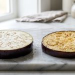 **Alt text:** Editorial food photography of two eggplant slices side-by-side on a marble counter. The left slice is raw, sprinkled with sea salt, and sweating moisture. The right slice is tender and pan-fried to a golden-brown.