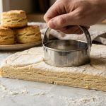 **Alt text:** Close-up of a metal biscuit cutter slicing through raw, laminated dough on a flour-dusted marble countertop, revealing flaky layers. Warm, blurry baked biscuits stand in the background.