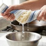 **Alt text:** A close-up of hands squeezing a white-and-blue striped kitchen towel filled with shredded potatoes, wringing starchy water into a stainless steel bowl on a bright marble countertop.