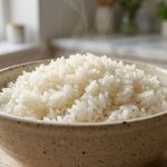 **Alt Text:** A close-up photograph of a ceramic bowl filled with fluffy, perfectly cooked white jasmine rice. Individual grains glisten under soft, natural side-light as a delicate wisp of steam rises, set against a softly blurred, minimalist kitchen background.