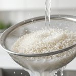 **Alt text:** Close-up of uncooked white jasmine rice being rinsed in a stainless steel colander under running water, with cloudy, starchy water draining out.