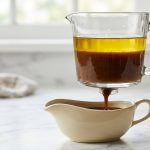 **Alt text:** A glass fat separator on a white marble countertop shows a layer of golden fat floating above a dark brown gravy, which pours from the bottom release valve into a ceramic gravy boat.