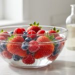 **Alt text:** A bright, close-up photograph of fresh strawberries, blueberries, and raspberries soaking in a clear glass bowl of water on a marble countertop. Tiny bubbles cling to the fruit, with a bottle of vinegar in the softly blurred background.