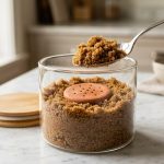 **Alt text:** A silver spoon scooping moist, golden-brown sugar from a glass canister on a marble countertop, with a round terracotta clay disk nestled in the sugar against a soft-focus, sunlit kitchen background.