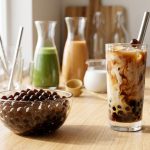 **Alt text:** A minimalist DIY bubble tea station bathed in warm morning light. In the foreground, a bowl of glossy tapioca pearls sits beside a tall glass of iced milk tea with marbled swirls. Carafes of matcha and tea are softly blurred in the background.