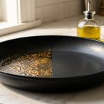 A close-up photograph of a black nonstick skillet on a white marble countertop, with its surface split to show one half covered in sticky, amber-colored baked-on residue and the other half pristine and smooth.