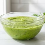 **Alt text:** A professional culinary photograph of a clear glass bowl filled with vibrant green guacamole on a white marble countertop. A thin, glistening layer of clear water rests flat on top of the guacamole as a protective seal, catching soft reflections of light. The background is softly blurred with a fresh lime half and cilantro sprigs under bright, natural window light.