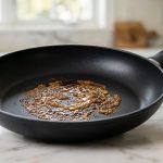 **Alt text:** A close-up of a matte-black nonstick skillet on a marble countertop, featuring a sticky, amber-colored patch of caramelized residue on its surface.