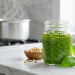 **Alt text:** A glass jar of vibrant green basil pesto on a marble countertop, surrounded by fresh basil leaves, pine nuts, and parmesan cheese, with a steaming pot in the blurry background.
