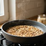**Alt text:** Close-up of rolled oats being dry-toasted to a golden brown in a modern ceramic pan. Soft morning light illuminates the crisp, textured oats as a gentle wisp of steam rises, with a blurred kitchen background.