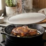 **Alt text:** A thick-cut ribeye steak with a golden-brown crust sears in a cast-iron skillet on a modern induction stovetop. A stainless steel splatter screen is lifted slightly, letting steam escape, while the surrounding countertop remains pristine.
