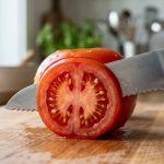 **Alt text:** Extreme close-up of a serrated steel knife making a clean, precise slice through a ripe red tomato on a light wooden cutting board, revealing its juicy interior and seeds.