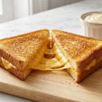 A perfectly golden-brown grilled cheese sandwich cut diagonally on a wooden board, with melted cheese pulling between the halves. In the soft-focus background, a ramekin of mayonnaise and a knife rest on a marble countertop.