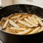 A close-up, high-angle photograph of hand-cut french fries cooking in bubbling golden oil inside a seasoned black cast-iron Dutch oven. Some fries are still pale and starchy, while others are starting to turn crisp and golden-brown at the edges.