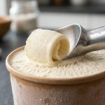 **Alt text:** A macro photograph of a brushed aluminum scoop carving a perfect curl of vanilla bean ice cream from a pint. Delicate frost crystals on the container's rim contrast with a glossy, slightly melted edge where the warm scoop cuts through, set on a dark slate countertop.