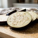 **Alt text:** Macro food photograph of raw eggplant slices on a wooden cutting board, sprinkled with coarse salt and sweating glistening beads of moisture.